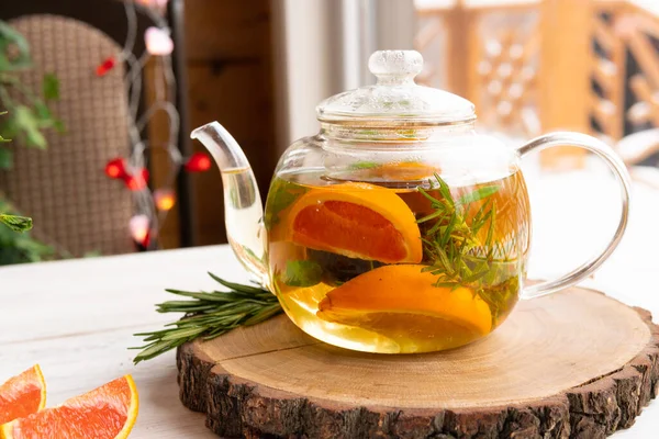 Healthy drink of Apple, orange and rosemary in a glass teapot on a wooden table by the window. Herbal tea is useful for health and prevention of diseases and viruses covid-19. — Stock Image Healthy Drink Apple Orange Rosemary Glass Teapot Wooden Table Window Stock Image