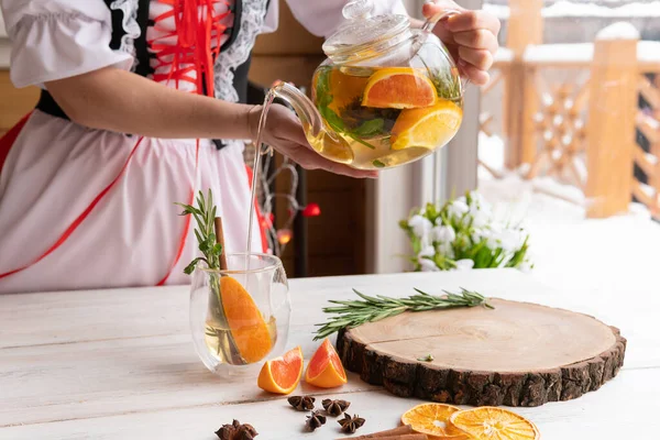 A woman drinks a healthy drink of Apple, orange and rosemary in a glass teapot on a wooden table by the window. Herbal tea is useful for health and prevention of diseases and viruses covid-19. — Stock Image Woman Drinks Healthy Drink Apple Orange Rosemary Glass Teapot Wooden Royalty Free Stock Images
