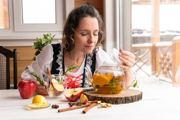 A woman drinks a healthy drink of Apple, orange and rosemary in a glass teapot on a wooden table by the window. Herbal tea is useful for health and prevention of diseases and viruses covid-19. — Stock Image Woman Drinks Healthy Drink Apple Orange Rosemary Glass Teapot Wooden Royalty Free Stock Photos