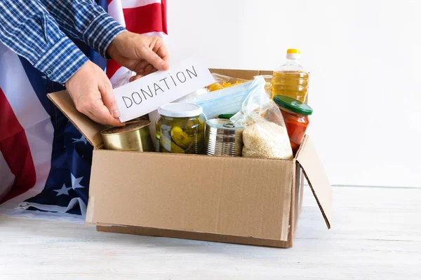 The hands are placed food. A donation box with various food items. Cardboard box with butter, canned food, cereals and pasta. U.S. flag.