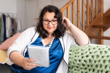 Koca vücutlu bir kadın kanepede oturuyor. Defterine bakıyor..