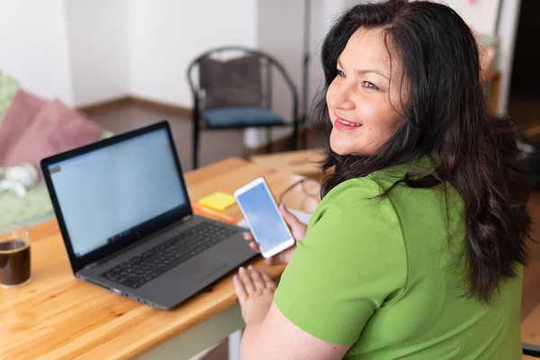 Fat woman sitting at a table at home, working with a laptop with a happy face. She\'s on the phone. Working on the Internet.