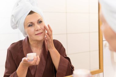 Kadınlar aynaya bakıp banyoda krem sürerler. Güzel bayan başını sarıp nemlendirici sürmüş..