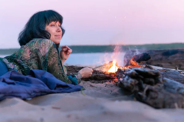 A woman sits by the fire in the evening on the shore of the lake. She looks thoughtfully