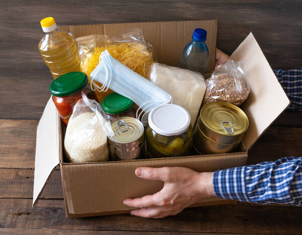 Картонная коробка с маслом, консервами, крупой и макаронами. Коробка для пожертвований с различными продуктами питания. Руки кладут в пищу.