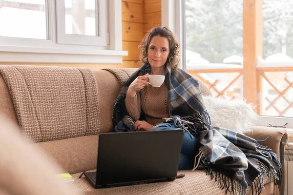 Woman with laptop working at home on the couch. She communicates on the Internet. She is holding a cup.