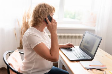 Orta yaşlı bir kadın ofiste dizüstü bilgisayarda çalışıyor. Telefonda konuşuyor..