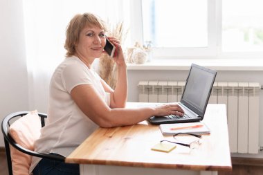 Orta yaşlı bir kadın ofiste dizüstü bilgisayarda çalışıyor. Telefonla konuşuyor..