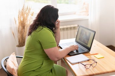 Şişman kadın, ofisteki pencerenin yanındaki masada dizüstü bilgisayarla çalışıyor. Akıllı telefonuyla konuşuyor..
