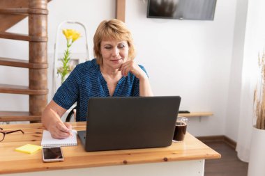 Masada çalışan orta yaşlı bir kadın laptop ekranına bakıyor. İnternette çalışıyor..
