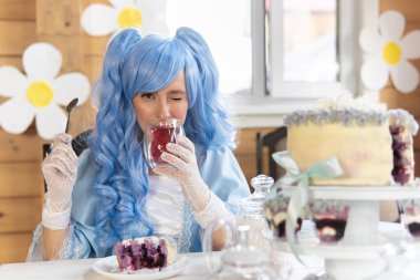 Masal kostümü giymiş mavi peruklu bir kadın çay içer ve sinsice gülümser.