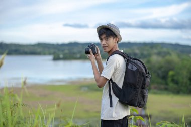 Sırt çantalı, dijital kameralı genç Asyalı gezgin kameraya güzel bir doğa gölü arka planıyla bakıyor. Seyahat tatil rahatlama konsepti