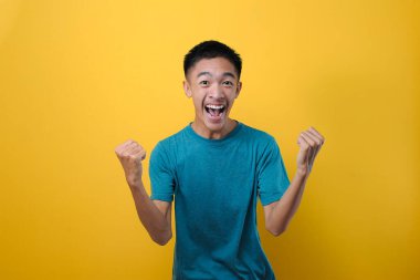 Mutlu Asyalı genç bir adamın portresi heyecanlı, iki elini kaldırıyor, sarı arka planda soyutlanmış.