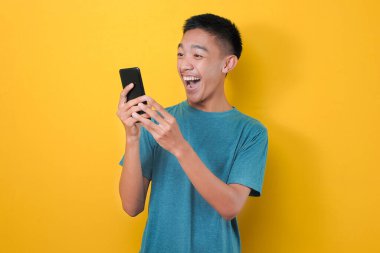 Sarı arka planda izole edilmiş, günlük mavi tişörtlü Asyalı adamın şok olmuş yüzü telefon ekranına bakıyor..