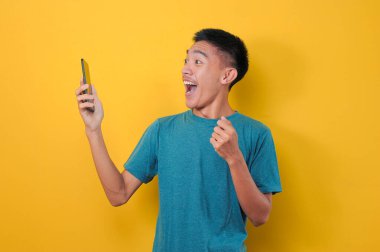 Sarı arka planda izole edilmiş, günlük mavi tişörtlü Asyalı adamın şok olmuş yüzü telefon ekranına bakıyor..