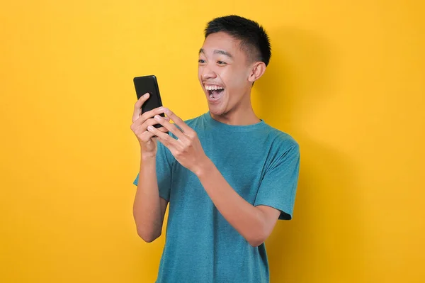 Sarı arka planda izole edilmiş, günlük mavi tişörtlü Asyalı adamın şok olmuş yüzü telefon ekranına bakıyor..