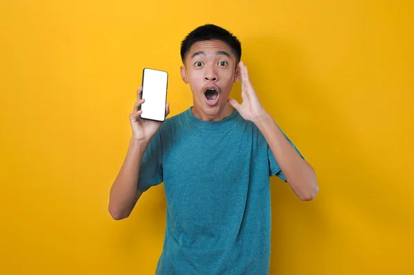 兴奋的年轻亚洲男子震惊地在摄像机前展示白色手机屏幕 被黄色背景隔离 — 图库照片