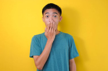 Şok olmuş genç Asyalı bir adam elini ağzını kapatıyor. Çok duygusal ve şaşırmış görünüyor. Sarı arka planda izole edilmiş.