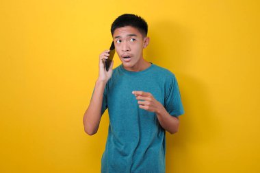 Şok suratlı genç Asyalı bir adam sarı arka planda izole edilmiş bir yabancıdan ya da dolandırıcıdan telefon almış.