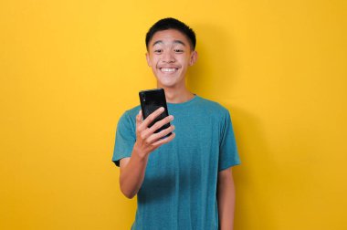 Akıllı telefonuyla gülümseyen ve telefon ekranına bakan mutlu Asyalı adam, online mağaza başvurusunda indirim alıyor, sarıda izole edilmiş.