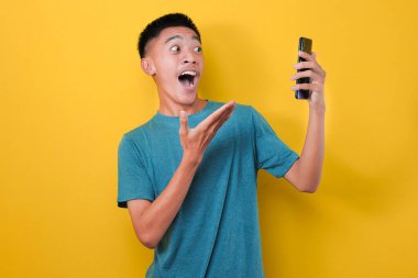Sarı arka planda izole edilmiş, günlük mavi tişörtlü Asyalı adamın şok olmuş yüzü telefon ekranına bakıyor..