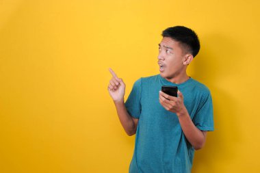 Elinde cep telefonu olan ve sarı arka planda izole edilmiş fotokopi alanına bakan, şok olmuş Asyalı genç adam.