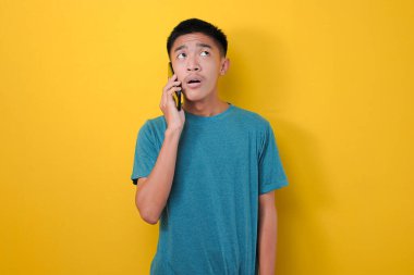 Şok suratlı genç Asyalı bir adam sarı arka planda izole edilmiş bir yabancıdan ya da dolandırıcıdan telefon almış.