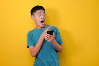 Elinde cep telefonu olan ve sarı arka planda izole edilmiş fotokopi alanına bakan, şok olmuş Asyalı genç adam.