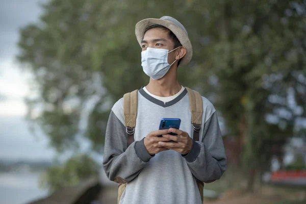 Asian Teenager man in sweater with protective face mask using smart phone. Asian man outside the river on the road uses a smartphone with mask face.