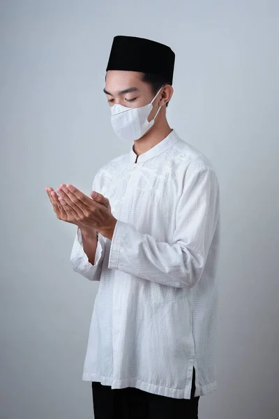 Asian Muslim Man wearing muslim clothes with medical mask and skull cap praying to God against on grey background