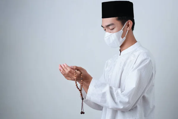 Asian Muslim Man wearing muslim clothes holding prayer beads with medical mask and skull cap praying to God against on grey background