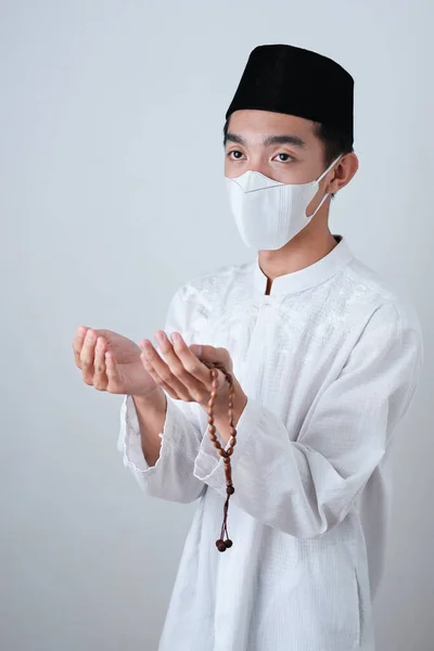 Asian Muslim Man wearing muslim clothes holding prayer beads with medical mask and skull cap praying to God against on grey background