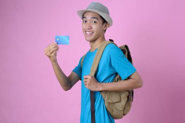 Kredi kartı tutan genç bir Asyalı turist. Genç gülümseyen Asyalı turist elinde kredi kartıyla seyahat etmeye hazır pembe arka planda.