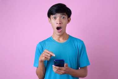 Şok olmuş genç Asyalı adam cep telefonunu, gözlerini ve ağzını pembe arka planda açık tutuyor.