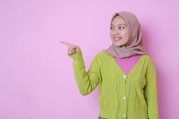 コピースペースの脇に魅力的な幸せな若いイスラム教徒のアジアのティーンエイジャーの笑顔ポイントは 広告がピンクの背景にイスラム教徒のカジュアルな服を着て示しています プロモーションコンセプト — ストック写真