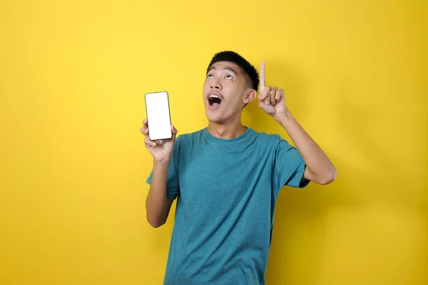 快乐的笑着的年轻亚洲男人 用另一只手拿着手机 用黄色的背景指着空旷的空间 — 图库照片