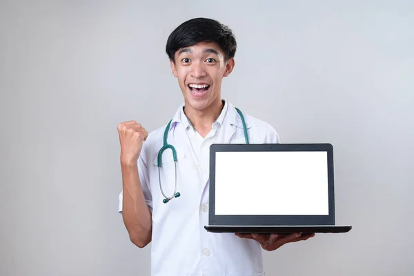 興奮したアジアの医師男がノートパソコンの応援を保持屈託のない興奮 灰色の背景に隔離された白い空白のノートパソコンの画面を示す — ストック写真