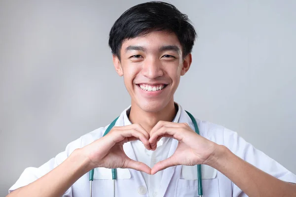 若いアジアの医師の男性の肖像灰色の背景に指で心臓の形のジェスチャーを示す — ストック写真