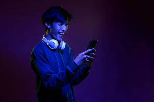 Portrait of excited handsome young asian man looking at the mobile phone wearing headphone in neon light. Futuristic neon lighting. Trendy style