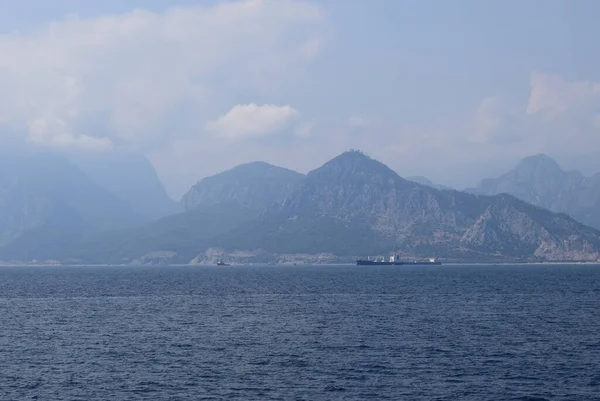 Cargo ship in calm blue Mediterranean in Anatalya. Traveling along the Mediterranean Sea, view of the rocky coast of Antalya. In the distance, cargo ships are visible.