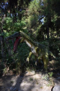 Kükreyen Baryonyx, arka plandaki orman parkında uzun otların üzerinde duran model dinozor heykeli. Türkiye 'de Dinopark, Goynuk. Tarih öncesi dinozor dişlek ağzını gösteriyor.