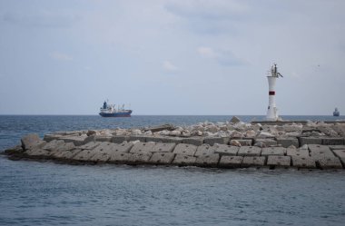 Limandan açık denize giden bir konteyner gemisi deniz fenerini geçiyor. Antalya Limanı, akşam güneşli ve bulutlu bir günde mavi gemi ve deniz feneri..