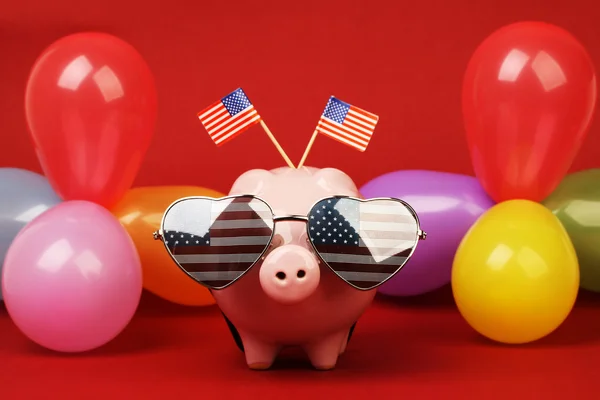 Piggy bank with retro sunglasses with USA flag and two small USA flags and balloons many colors on red background — Stock Photo, Image