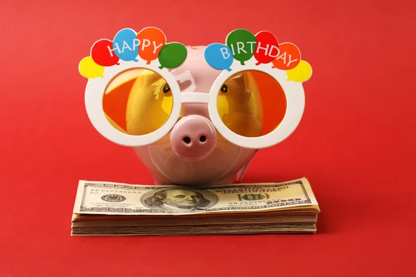 Hucha con gafas de fiesta de cumpleaños feliz de pie en la pila de dinero billetes de cien dólares americanos sobre fondo rojo — Foto de Stock