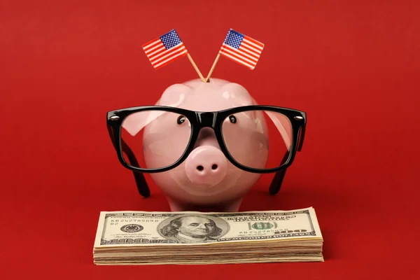 Tirelire avec monture de lunettes noire et deux petits drapeaux des États-Unis debout sur pile de billets de cent dollars américains sur fond rouge Photos De Stock Libres De Droits