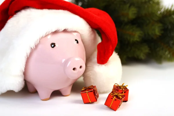 Part of piggy bank with Santa Claus hat and three little gifts and christmas tree standing on white background — Stock Photo, Image