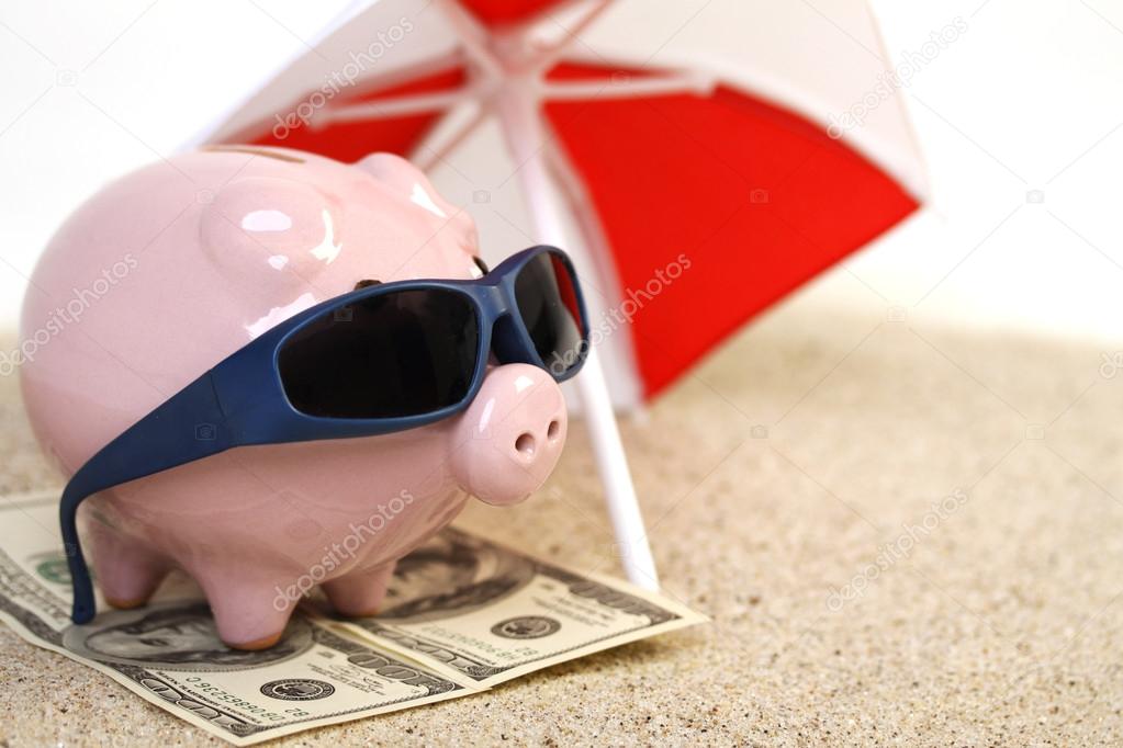 Summer piggy bank standing on towel from greenback hundred dollars with sunglasses on the beach sand unter red and white sunshade