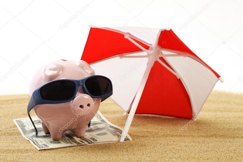 Summer piggy bank standing on towel from greenback hundred dollars with sunglasses on the beach sand under red and white sunshade