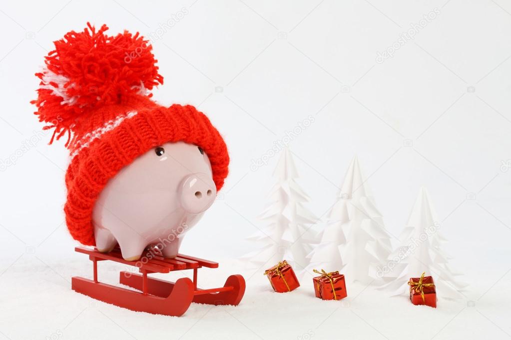 Piggy box with red hat with pompom standing on red sled on snow and around are snowbound trees and three gifts with gold bow