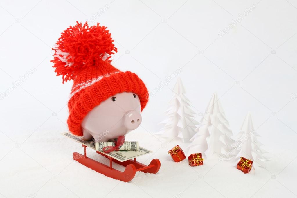 Piggy box with red hat with pompom standing on red sled with blanket from greenback hundred dollars on snow and around are snowbound trees and three gifts with gold bow